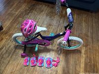 Side view of Huffy Glitter kids bicycle with pink helmet mounted on seat and set of pink protective gear on the floor