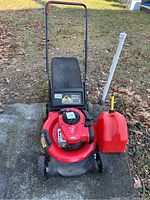 Full view of red Craftsman lawn mower with attached black grass catcher and a red gasoline container on a concrete slab surrounded by autumn leaves.