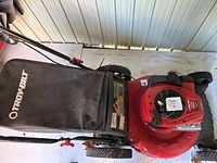 Photo showing the top and side view of a red Troy-Bilt lawn mower with black grass catcher bag, stored indoors on a concrete floor.