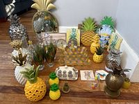 Overview of various pineapple-themed decorative items including glasses, figurines, trays, and mirrors arranged on floor.