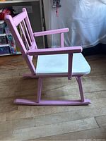 Side view of mini wooden rocking chair showing pink arms, backrest with vertical slats, white seat, and curved rockers on floor.