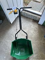 Seed spreader from handle view showing black metal frame, yellow adjustment lever and green plastic hopper.