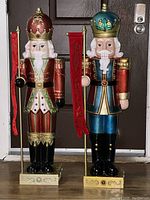 Photo showing two tall plastic nutcracker figures, one red and cream with gold and green accents holding a spear, the other blue and black with gold accents holding a red flag. Both are lit and standing on decorative bases indoors.