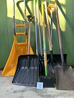 Photo showing all eight items: three snow shovels (one yellow plastic with curved handle, two black plastic), black metal square point shovel, two green-handled garden tools (small shovel and hoe), yellow plastic ice scraper, and a small garden shovel with wooden handle.