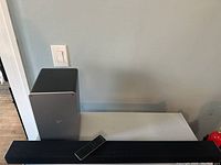 Main soundbar speaker, subwoofer, and remote control shown on a white surface near a wall