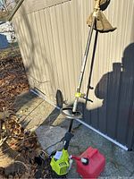 Full view of Ryobi gas trimmer with shaft, handle, guard and part of the fuel tank base visible, shown leaning against shed