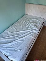 Twin bed frame with white upholstered headboard shown from a side angle with mattress on it.