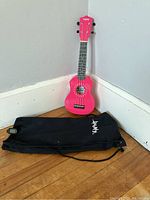 Pink Vanphy kid's ukulele standing against the wall next to a black Vanphy-branded carrying bag on a wooden floor.
