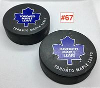 Two black hockey pucks with Toronto Maple Leafs blue maple leaf logos and white text sitting on white background.