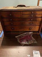 Front view of the antique wooden tool chest showing multiple small drawers and a plastic bag with small items on the table.