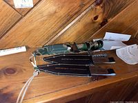 Set of three throwing knives with black leather sheaths on wooden surface. Two knives have ornamental silver handles; one has a camo green handle.