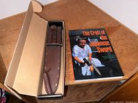 Photo showing artisan Japanese sword in dark brown leather sheath inside an open cardboard box, with the book "The Craft of the Japanese Sword" beside it on a wooden table.