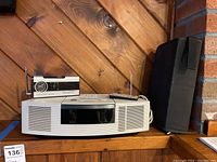 Front view of Bose Wave Radio CD player, transistor clock radio, and black speaker case on wooden shelf against wood panel wall
