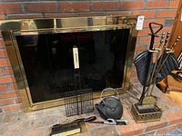 Photo showing fireplace with brass framed screen, fireplace tool set with shovel, poker, brush in ornate stand, black metal kettle, Char-Broil grilling basket, brass brush and pan, metal lighter.