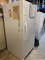 White Electrolux upright freezer shown from a left angled front view in basement, with note and decorations on the door.