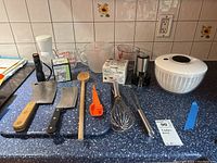 Wide shot of all kitchenware items on blue countertop including cleavers, spoons, whisks, measuring cups, salad spinner, and other kitchen tools.