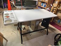 White laminated sewing table with black metal legs and attached white plastic storage tray with compartments.