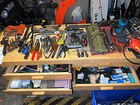 Wide view of assortment of hand tools and accessories laid out on wooden surface with drawers below, showing many screwdrivers, pliers, wrenches, hammers and miscellaneous small tools
