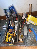 Photo of open toolbox packed with assorted hand tools such as saw, drills, screwdriver, hammer and various hardware items on carpeted floor.