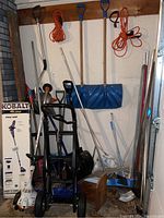 Photo showing overall collection of yard tools including snow shovels, dolly, and other tools against wall.