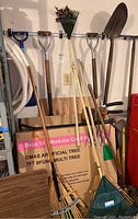 Photo showing assorted yard tools hanging on a rack including multiple shovels, rakes, and an axe. A boxed artificial Christmas tree is visible on the floor behind the tools.