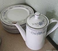 Coffee pot with lid and stacked dinner plates in background highlighting matching floral motif and silver trim