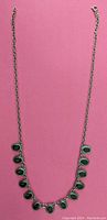 Full view of the necklace laid flat on a pink background showing the silver-tone chain with black oval accents arranged evenly across the front.