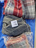 Three small throw blankets: one red and gray plaid, one gray with lighter plaid pattern, one red and cream wrapped in plastic.