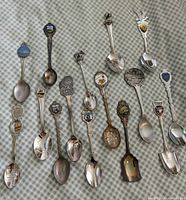 Photo of fifteen assorted vintage souvenir collector spoons laid out on cloth with various decorative handles from locations such as St. Martins, Banff, Nassau, EPCOT Center, showing enamel and metal designs.
