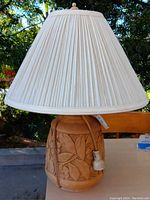 Table lamp with carved floral base and cream pleated lampshade, photographed outdoors to show design and condition.
