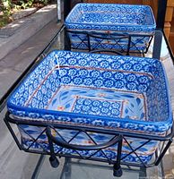 Two ceramic baking dishes with blue and white floral patterns sitting in black metal wire holders placed outdoors.
