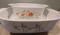 Photo showing both vintage CorningWare dishes, rectangular dish with purple iris flowers and square dish with multicolor flowers inside the larger dish.
