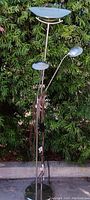 Full view of the chrome floor lamp with adjustable arms and stationary center light, shown outdoors against green foliage.