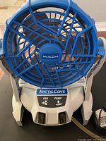 Front view of blue and white Arctic Cove battery operated fan, showing fan grill, handle, and control panel with FAN and PUMP switches.