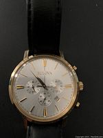 Front view of gold-tone Bulova chronograph watch with silver dial, black leather strap, three subdials, and date window.