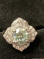 Clear photo of the ring showing the central mossanite stone surrounded by clear stones arranged in a symmetrical floral pattern on a silver-tone setting, placed on a black textured surface.