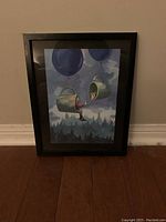 Full view of framed artwork showing two green mugs suspended by blue balloons with a person and dog inside each mug against a cloudy sky and forest backdrop.