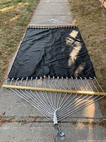 Full view of the black fabric hammock laid flat on a sidewalk, showing wooden spreader bars and white ropes attached to metal ring.