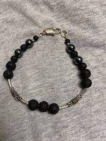 Full view of sterling silver bracelet featuring black lava stones, polished beads, silver tubes, and lobster claw clasp on grey fabric background.