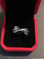 Front angle view of blue-green moissanite ring inside a red velvet jewelry box, showing the crossover band and two stones.