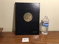 Front view of a large black hardcover book with a gold emblem and embossed name on the cover, placed upright next to a water bottle for scale.