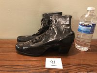 Side view of black leather antique ladies ankle boot with mid-height heel and several eyelets and hooks for lacing, displayed next to a water bottle for size reference.