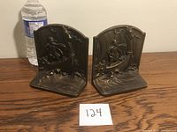 Two cast bronze bookends with relief Viking scenes on ships, shown side by side on wooden surface with water bottle for scale.