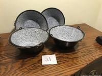 Set of four enamelware kettles shown with white speckled enamel interior and black exterior with wire handles on a wooden table