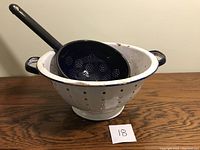 Two kitchen strainers on wood table: large white enamelware colander and smaller blue enamelware strainer with handle.