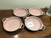 Four pink enamelware serving bowls with black rims and two handles each, placed on a wooden surface.