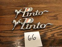 Photo of two Ford Pinto vehicle emblems on a wooden surface with a tag marked '66'.