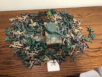 A large pile of assorted vintage style plastic toy soldiers in green and tan colors along with a camouflaged plastic tank toy on a wooden surface.