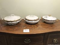 Three covered vegetable dishes placed side by side on a wooden table with lids on, showing decorative edges and handles.