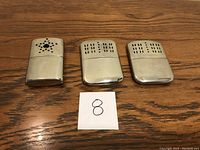 Photo showing three vintage metal butane hand warmers on wooden surface with paper marked '8'